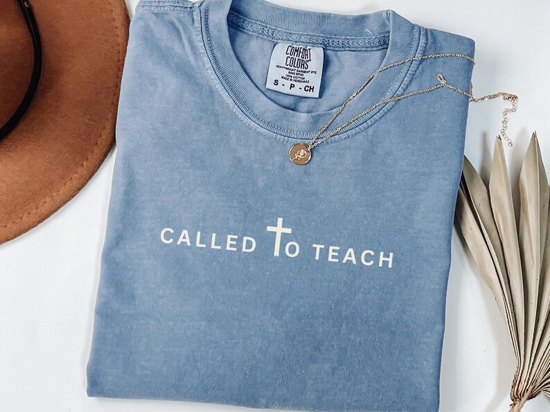 May include: A light blue t-shirt with the text "CALLED TO TEACH" and a white cross in the centre. The shirt is made of a soft, comfortable fabric and has a distressed look.