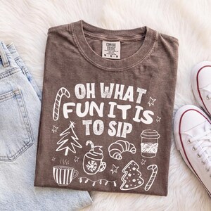 May include: A brown short-sleeved t-shirt featuring white graphic illustrations of a Christmas tree, a coffee cup, a croissant, and a candy cane. The text reads "OH WHAT FUN IT IS TO SIP" in a bold, white, distressed font style.