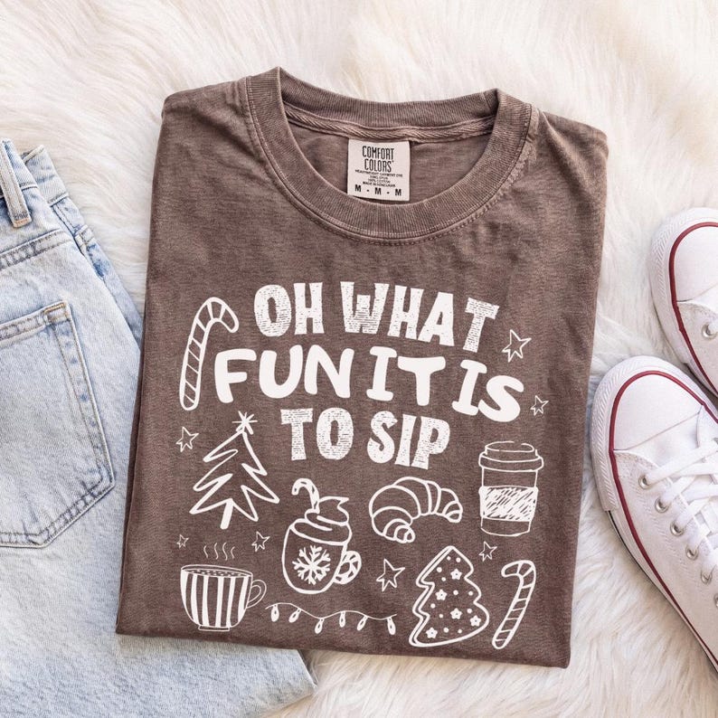 May include: A brown short-sleeved t-shirt featuring white graphic illustrations of a Christmas tree, a coffee cup, a croissant, and a candy cane. The text reads "OH WHAT FUN IT IS TO SIP" in a bold, white, distressed font style.