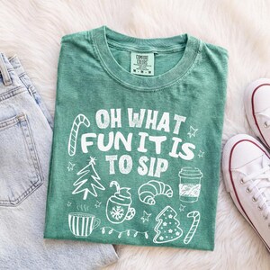 May include: A green short-sleeved t-shirt featuring white graphic illustrations of a candy cane, a Christmas tree, a mug, a croissant, and a coffee cup, with the text "OH WHAT FUN IT IS TO SIP" printed in a bold, distressed white font.