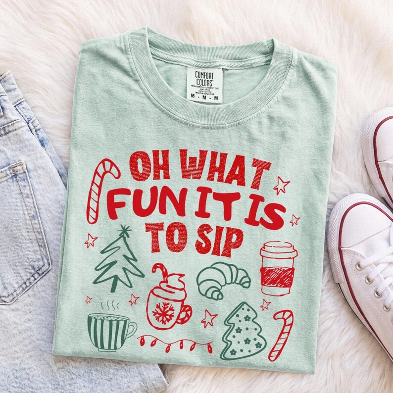 May include: A light green t-shirt featuring red text that reads OH WHAT FUN IT IS TO SIP. The design includes red line art illustrations of a candy cane, a Christmas tree, a mug with a snowflake, a croissant, a coffee cup, and a star.
