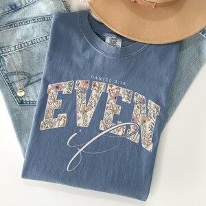 Puede incluir: Una camiseta azul polvoriento con la palabra "EVEN" en un dise&ntilde;o floral y el texto "Daniel 3:18". Tambi&eacute;n se ven un sombrero marr&oacute;n y un collar dorado. La camiseta est&aacute; doblada sobre un par de vaqueros azul claro.