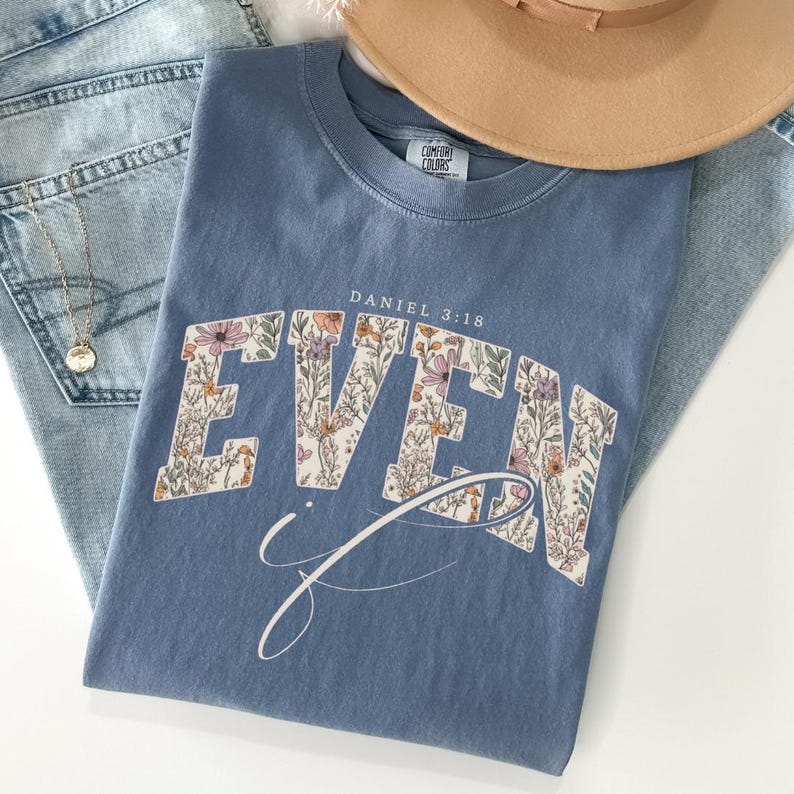 Puede incluir: Una camiseta azul polvoriento con la palabra "EVEN" en un dise&ntilde;o floral y el texto "Daniel 3:18". Tambi&eacute;n se ven un sombrero marr&oacute;n y un collar dorado. La camiseta est&aacute; doblada sobre un par de vaqueros azul claro.