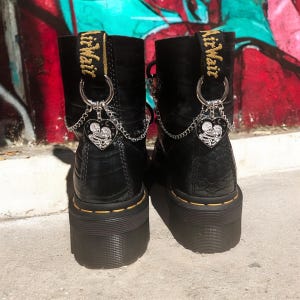 Puede incluir: Botas de plataforma de cuero negro con cadenas plateadas y dijes en forma de corazón que dicen "Airwair" en la parte posterior.
