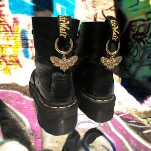 Puede incluir: Botas de plataforma de cuero negro con abalorios de abeja dorada atados a los cordones. Los abalorios tienen el texto "Air Mail".