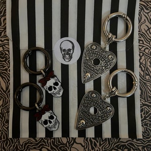 Könnte beinhalten: Ein Set aus vier Schlüsselanhängern mit einem schwarz-weißen Totenkopf mit rotem Bandana, einem silbernen herzförmigen Ouija-Brett und einem silbernen Kreis-Schlüsselanhänger. Die Schlüsselanhänger sind auf einem schwarz-weiß gestreiften Hintergrund abgebildet.
