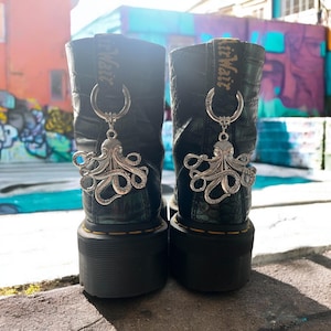 May include: Two black platform boots with silver octopus charms. The boots have a textured leather effect and a thick, ridged sole. The charms are attached with a silver ring. The boots are approximately 20 cm high.