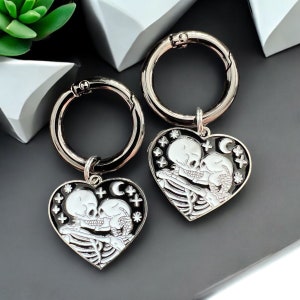 May include: Two silver keyrings with black heart-shaped charms featuring a white skeleton couple kissing. The charms are decorated with white stars and a crescent moon.