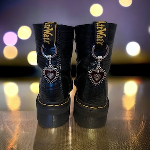 Peut inclure: Bottes en cuir noir avec une étiquette jaune "AirWair" sur chaque botte. Chaque botte a un charme argenté en forme de cœur avec un centre rouge et un motif d'œil argenté.