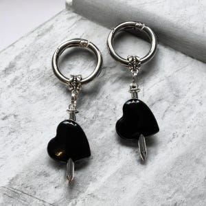 May include: A pair of silver-toned hoop earrings with black heart-shaped pendants. Each earring features a small decorative element connecting the hoop to the heart. The heart pendants have a glossy finish and a silver-toned spike at the bottom.