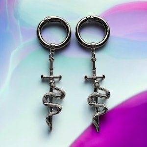 May include: A pair of silver earrings with a snake wrapped around a sword charm. The earrings are made of metal and have a circular shape.