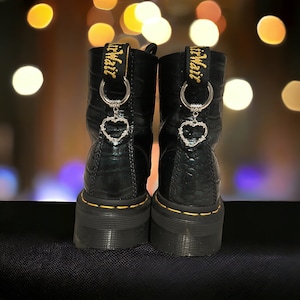 Könnte beinhalten: Schwarze Lederstiefel mit gelben Nähten und einem herzförmigen Anhänger an jedem Stiefel. Die Anhänger sind silberfarben mit einem Stacheldraht-Design. Die Stiefel haben ein Dr. Martens Logo auf der Rückseite.
