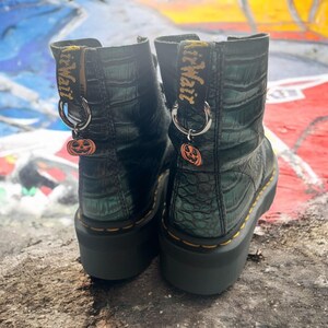 May include: Two black platform boots with a crocodile skin effect. Each boot features a yellow pull tab with "Air Wair" printed on it. Silver hoops hang from the tabs, each with an orange pumpkin charm. The boots have yellow stitching.