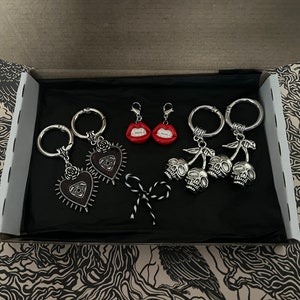 Op de afbeelding: Een set van vijf sleutelhangers in een zwarte doos. Twee sleutelhangers hebben zilveren harten met een oogontwerp. Twee sleutelhangers hebben zilveren kersen met schedelcharms. Eén sleutelhanger heeft een rode, open mond met tanden.