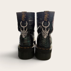 May include: A pair of black boots with a crocodile-embossed texture and a green iridescent sheen. Each boot features a black heel loop with the text AirWair, a silver metal ring, and a dangling silver-toned demon mask charm on the back.