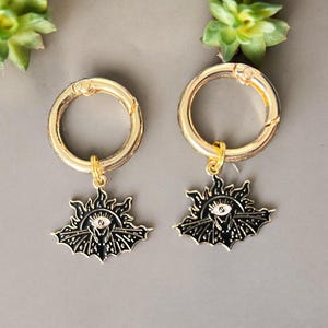 May include: A pair of gold hoop earrings with a black and white sun and eye charm hanging from each hoop.