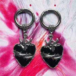 May include: Two silver hoop earrings with black heart charms. The hearts are detailed with a cross, thorns, and a black enamel finish.