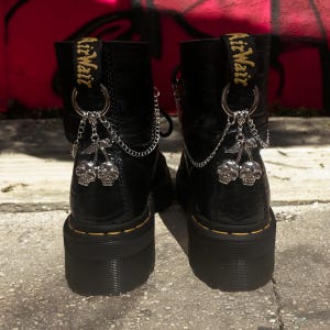 Könnte beinhalten: Schwarze Lederstiefel mit silbernen Totenkopf- und Kirsch-Anhängern, die an Ketten befestigt sind. Die Stiefel haben eine dicke Plateau-Sohle und der Text "AirWair" ist auf der Rückseite jedes Stiefels sichtbar.
