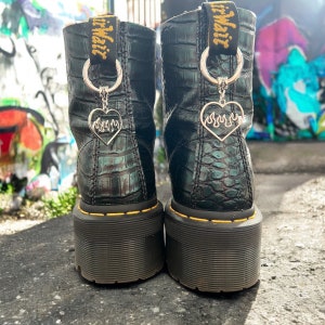 May include: Black leather boots with a platform sole and a yellow stitching detail. The boots have a silver charm attached to each boot with a heart design and flames.