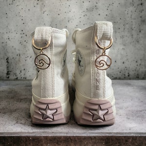 Könnte beinhalten: Paar cremefarbene High-Top-Sneaker mit Sternmuster auf der Sohle und einem goldfarbenen Wellenanhänger. Die Sohlen sind hellrosa. Auf der Rückseite der Sneaker steht das Wort "ONE".