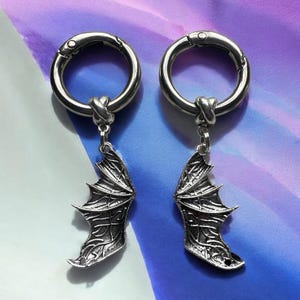 May include: A pair of silver metal earrings with a dragon wing charm hanging from each. The earrings are made of a thick metal ring with a clasp closure.