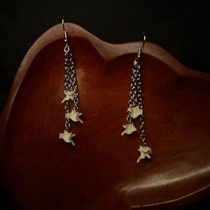 Peut inclure: Une paire de boucles d'oreilles en chaîne argentée avec des breloques en os blanc qui pendent. Les boucles d'oreilles sont présentées sur une surface en bois.