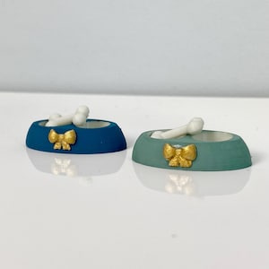 May include: Two miniature pet food bowls, one teal and one blue, each with a gold bow and a white bone inside. The bowls are small, likely for a dog or cat, and sit on a white surface.