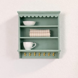Peut inclure: Étagère murale vert d'eau avec trois niveaux et un bord supérieur festonné. Elle contient un bol blanc, du tissu plié et une tasse blanche. Sept crochets dorés sont en dessous. L'étagère est fixée sur un mur blanc. Mesure environ 40 cm de large.