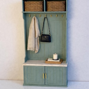 May include: A sage green hallway storage unit featuring a bench with two lower cabinet doors, a vertical panelled backboard with seven gold hooks, and an upper shelf holding two woven baskets. A beige scarf and black purse hang from the hooks.