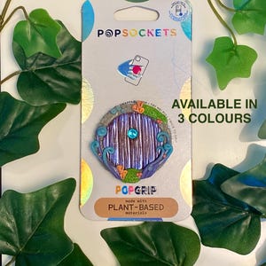 May include: A PopSockets PopGrip featuring a fantasy door design with a blue gem. The packaging reads "POPSOCKETS" and "AVAILABLE IN 3 COLOURS". The PopGrip is made with plant-based materials.
