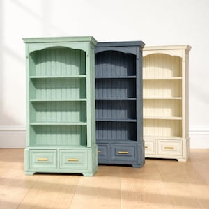 May include: Three miniature wooden bookcases standing side by side, each featuring three open shelves and two small drawers at the base with gold-toned horizontal handles. The units are painted in sage green, dark navy blue, and cream white.