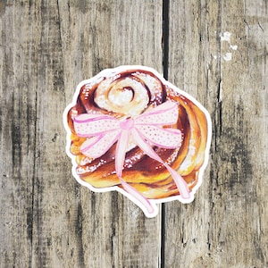 Pode incluir: Um autocolante com uma ilustração em aquarela de um rolo de canela. A pastelaria é de cor castanha quente, polvilhada com açúcar de confeiteiro e adornada com um laço rosa com bolinhas brancas. O autocolante está sobre um fundo de madeira.