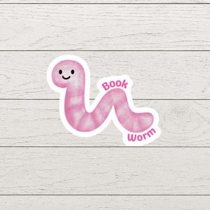 May include: A pink sticker featuring a smiling cartoon bookworm with the words "Book Worm" in a playful font. The worm has a segmented body and is set against a white background.