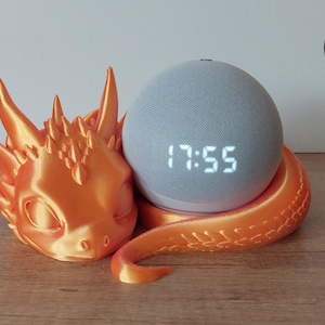 Könnte beinhalten: Orangefarbener 3D-gedruckter Drache, der einen grauen intelligenten Lautsprecher mit einer Digitaluhr hält, die 17:55 anzeigt.
