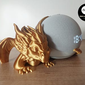 Könnte beinhalten: Eine goldfarbene 3D-gedruckte Drachenfigur, die einen grauen Amazon Echo Dot Smart Speaker mit einer Digitaluhr hält, die 18:48 anzeigt. Der Drache hat Flügel und ein detailliertes Gesicht.
