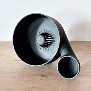Könnte beinhalten: Ein schwarzes, 3D-gedrucktes Turbinengehäuse mit Spiraldesign. Das Gehäuse hat einen großen, kreisförmigen Einlass und einen kleineren, gebogenen Auslass. Das Innere hat detaillierte Lüfterblätter. Das Objekt wird auf einer Holzoberfläche präsentiert.