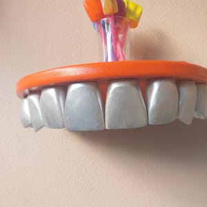 Puede incluir: Un estante de dientes de plata con un borde naranja. El estante está montado en una pared y tiene un pequeño jarrón de vidrio con flores de plástico de colores en la parte superior.