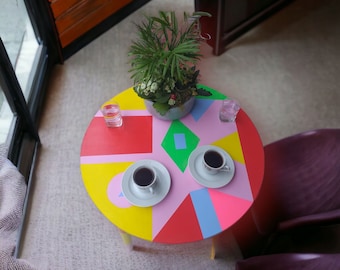 Könnte beinhalten: Ein runder, bunter Tisch mit geometrischen Formen in Rosa, Gelb, Rot, Blau und Grün. Zwei Tassen Kaffee und zwei Gläser Wasser stehen auf dem Tisch. Im Hintergrund steht eine Topfpflanze.