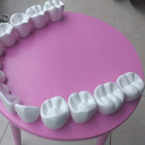 Puede incluir: Una mesa rosa caprichosa con dientes de cerámica blanca dispuestos en el borde. La mesa tiene tres patas rosas y una tapa redonda. Los dientes son de varios tamaños y formas.