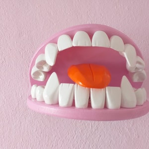 Può includere: Una scultura stravagante a forma di bocca aperta, dipinta di rosa, con grandi denti bianchi e una lingua arancione. La scultura è montata su un muro rosa, creando un'esposizione giocosa.