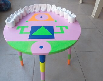 May include: A pink round table with green, blue, and orange geometric shapes painted on the top. The table has three legs painted in pink, yellow, blue, and green.