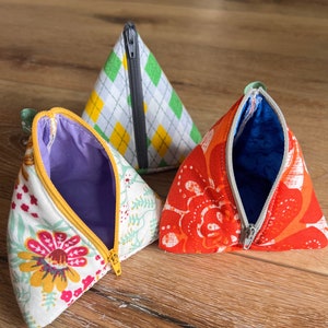 May include: Three triangular zippered pouches with different fabric patterns. The first pouch is white with a floral pattern, the second is green and white with a plaid pattern, and the third is orange with a floral pattern.