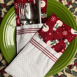 May include: Two red and white fabric place card holders with a snowman pattern. The holders have a white pocket with red stitching and a white snowflake pattern. A silver fork and spoon are in one of the holders.