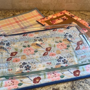 May include: A clear glass baking dish sits on a quilted hot pad with a floral pattern. The hot pad has a blue background with pink, red, and yellow flowers and birds. The hot pad has a blue border.