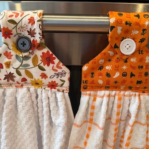 May include: Two hanging kitchen towels with fabric toppers. The left towel has a white topper with autumn leaves, mushrooms, and a blue button. The right towel has an orange topper with Halloween icons, the number 31, and a white button.