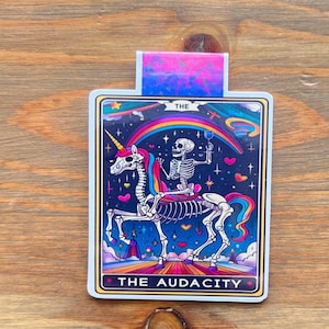 May include: A rectangular tarot card featuring a skeleton riding a unicorn under a rainbow. The card has the words "THE AUDACITY" at the bottom and "THE" at the top. The unicorn has a rainbow mane and tail, and the skeleton is holding a glass.