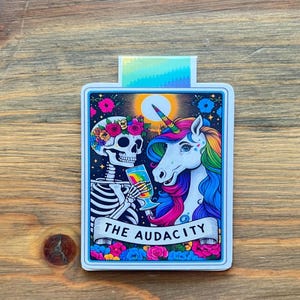 May include: A rectangular sticker featuring a skeleton and a unicorn. The skeleton wears a floral crown and holds a rainbow-colored glass. The unicorn has a rainbow mane and horn. The text "THE AUDACITY" is on a banner below the figures. The sticker has a colorful border.