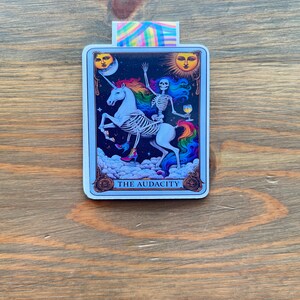 May include: A rectangular sticker featuring a tarot card design. The card depicts a skeleton riding a unicorn with a rainbow mane and tail. The card has a blue border with the words "THE AUDACITY" at the bottom. The background is a wooden surface.
