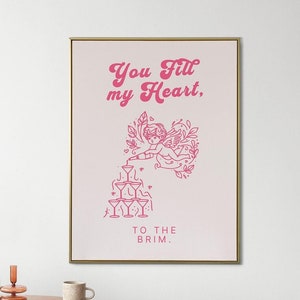 May include: Pink and white printable wall art with a gold frame. The art features a pink illustration of a cupid pouring champagne into a pyramid of glasses. The text reads "You fill my heart, to the brim."