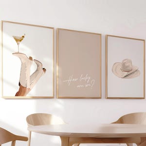 Puede incluir: Tres impresiones artísticas enmarcadas con marcos dorados. Las impresiones presentan un cóctel, botas de vaquero, un sombrero y el texto "How lucky are we?" El arte está en tonos neutros, adecuado para la decoración del hogar.